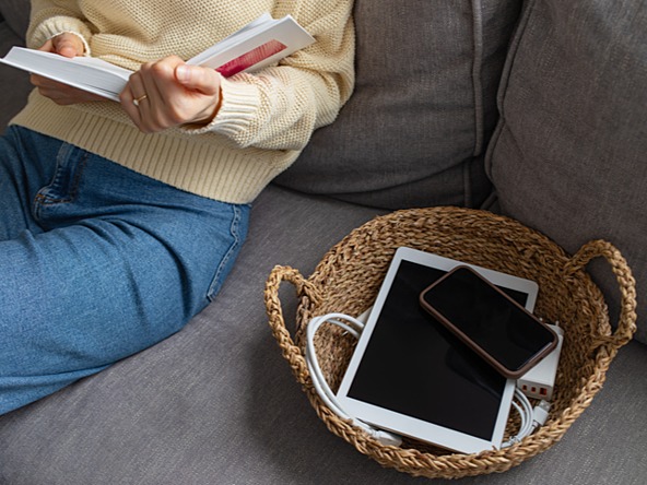 person reading a book on couch next to basket containing switched-off phone and tablet